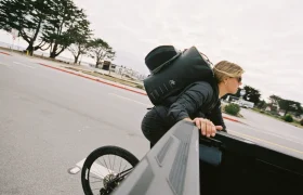 Una persona con una gran mochila negra se inclina sobre el lateral de una camioneta, sujetando el borde, mientras destaca una rueda de bicicleta -perfecta para los ciclistas que confían en ASSOS x Db para su sistema de viaje-; los árboles se balancean al fondo en un día nublado.