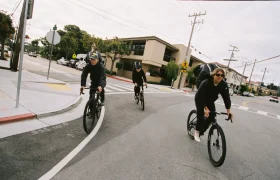 Tres ciclistas con ropa oscura y cascos circulan por una calle de la ciudad, doblando una esquina cerca de una intersección. La escena capta la energía urbana, encarnando el espíritu de aventura de ASSOS x Db y un innovador sistema de viaje.
