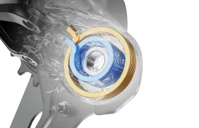 Primer plano de un componente mecánico Shimano DEORE XT M8200 con una carcasa transparente, que deja ver una bobina circular azul y dorada en el interior de la estructura metálica, diseñada para prolongar la vida útil del cable.