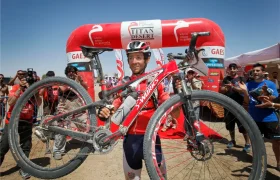 Un ciclista con ropa de carrera sostiene una bicicleta de montaña frente al arco inflable rojo Alpinum TotalEnergies en la línea de meta, rodeado de gente que lo anima y fotógrafos.