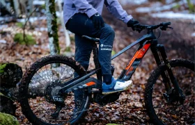 Una persona vestida de ciclista con una bicicleta de montaña Rotwild R.EXC embarrada en un sendero cubierto de hojas en una zona boscosa, lista para competir incluso en condiciones difíciles.