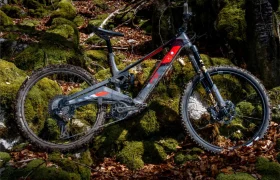 Una bicicleta de montaña Rotwild R.EXC de suspensión total con barro en los neumáticos está aparcada sobre rocas musgosas y hojas caídas en una zona boscosa, lista para competir en cualquier terreno.