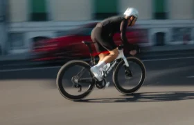 Un ciclista, vestido con ropa negra y casco blanco, recorre a gran velocidad las calles de Nevers montado en una bicicleta de carretera LOOK 795 Blade RS 3, con un coche rojo difuminado al fondo.