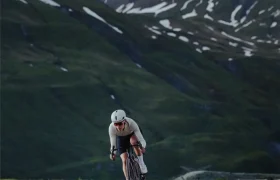 Un ciclista que monta una LOOK 795 Blade RS 3, con casco y equipación, desciende por una sinuosa carretera de montaña cerca de Nevers, con colinas verdes y un pico cubierto de nieve al fondo.