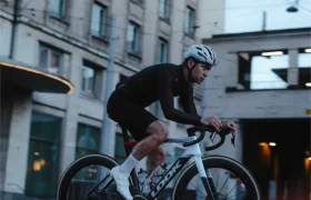 Un ciclista, que lleva casco y ropa de ciclismo negra, circula en una bicicleta de carretera LOOK 795 Blade RS 3 por una calle de la ciudad, frente a un gran edificio, al atardecer.