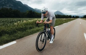 Un ciclista, equipado con casco y ropa de ciclismo, circula en una LOOK 795 Blade RS 3 por una carretera rural asfaltada en Nevers, con las montañas y la vegetación como telón de fondo.
