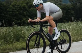 Un ciclista, con casco blanco y equipación de ciclismo, circula en una bicicleta de carretera LOOK 795 Blade RS 3 negra por una carretera asfaltada cerca de Nevers, con montañas verdes y árboles al fondo.