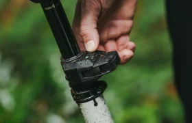 Una mano ajusta la palanca de la tija del asiento Tija RockShox Reverb AXS, cubierta de suciedad y gotas de agua, sobre un fondo verde borroso, perfecto para los entusiastas del MTB que buscan un rendimiento de vanguardia.