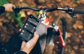 Una persona sostiene un smartphone con una aplicación de ciclismo mientras está sentada en una bicicleta de montaña roja en un bosque sereno. La MTB está equipada con funciones avanzadas como la RockShox Reverb AXS, que integra la tecnología de vanguardia Air-Over-Air para una experiencia de conducción mejorada.