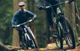 Dos ciclistas recorren una pista forestal en bicicleta de montaña, con sus ropas oscuras mimetizándose con el frondoso entorno. Equipados con la tecnología RockShox Reverb AXS, dominan cada curva con precisión, garantizando la seguridad con sus cómodos cascos.