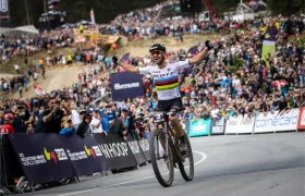 Un ciclista cruza la línea de meta con los brazos en alto en señal de victoria mientras una gran multitud le aclama en una carrera profesional de bicicleta de montaña, en honor a la retirada del legendario Nino Schurter.