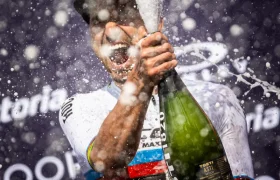 Un ciclista con un maillot de rayas arco iris rocía champán con una amplia sonrisa en la etapa, celebrando la retirada de Nino Schurter mientras las gotas y la espuma llenan el aire.