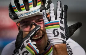 Un ciclista con casco y guantes de rayas arco iris sonríe y gesticula con las manos, mostrando con orgullo los guantes y su rostro, un guiño al célebre legado del retirado Nino Schurter.