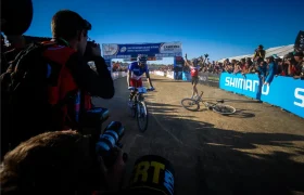Un ciclista cruza la línea de meta mientras otro -posiblemente en señal de retirada de Nino Schurter- yace en el suelo, rodeado de fotógrafos y espectadores.
