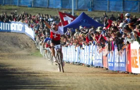 Un ciclista con un maillot suizo -que celebra la retirada de Nino Schurter- levanta un brazo en señal de victoria mientras cruza la línea de meta, animado por una multitud detrás de unas barricadas en una prueba ciclista.
