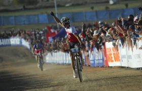 Un ciclista ataviado con un maillot blanco y rojo levanta un brazo en señal de victoria al cruzar la línea de meta, haciéndose eco de momentos como la retirada de Nino Schurter, con espectadores animando a lo largo del recorrido.