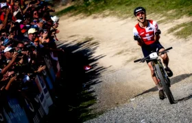 Un ciclista con maillot rojo y negro celebra al cruzar la línea de meta, evocando recuerdos del retirado Nino Schurter, mientras una multitud de espectadores anima en el lado izquierdo de una pista de tierra.