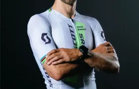 Un ciclista con un maillot blanco con los logotipos de los patrocinadores, de pie, con los brazos cruzados, sonriendo, y con una gorra negra sobre un fondo oscuro, captura el momento de la retirada de Nino Schurter.