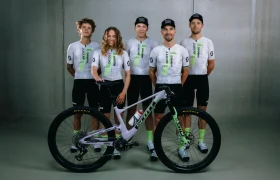 Cinco ciclistas con uniformes a juego se colocan detrás de una bicicleta de montaña, posando para una foto de grupo sobre un fondo liso de interior, celebrando la retirada de Nino Schurter.