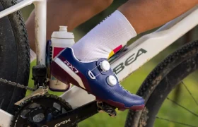 Primer plano del pie de un ciclista que lleva unas zapatillas Shimano azules en un pedal; se aprecian el cuadro blanco de la bicicleta, un bidón y unos calcetines blancos. Perfectas para los ciclistas de gravel que buscan tecnología de competición.