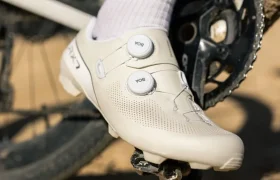 Primer plano de una persona que lleva unas zapatillas blancas de ciclismo Shimano RX710 enganchadas al pedal de una bicicleta, con parte del juego de bielas de la bicicleta y un camino de tierra visibles al fondo, lo que pone de relieve la tecnología Gravel para un rendimiento óptimo en terrenos accidentados.