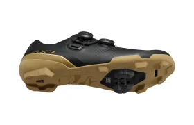 Se muestra una zapatilla de ciclismo negra con suela de color beige vista desde el lateral y desde abajo, que cuenta con una cala, una suela con tacos y un sistema de cierre de doble dial. Diseñadas para ciclistas de gravel, estas zapatillas Shimano incorporan tecnología de competición para ofrecer un rendimiento excepcional en terrenos variados.