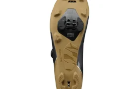 Primer plano de la suela de una zapatilla de ciclismo Shimano con dibujo para grava de color beige, cala negra y logotipo visible, fotografiada sobre un fondo blanco. Este diseño incorpora tecnología de competición ideal para salidas exigentes.