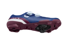 Unas zapatillas de ciclismo Shimano de color azul y granate, mostradas de perfil y desde abajo, con dos ruedas de ajuste blancas, una cala negra fijada a la suela y tecnología de competición ideal para aventuras sobre grava.