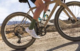 Una persona conduce una bicicleta de grava de color canela por un camino de tierra en un paisaje montañoso y desértico.