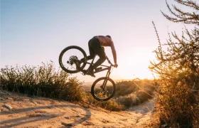 Una persona monta en bicicleta de montaña con cambio mecánico Shimano DEORE XT M8200 al saltar desde un montículo de tierra en un sendero al atardecer, rodeada de matorrales y vegetación seca, garantizando una larga vida al cable en cada emocionante recorrido.