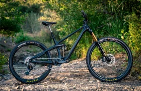 Una bicicleta de montaña de doble suspensión con cuadro negro, cambio mecánico Shimano DEORE XT M8200 y neumáticos de tacos se encuentra en un camino de tierra rocoso rodeado de frondosa vegetación. «Larga vida al cable» en cada aventura extrema.