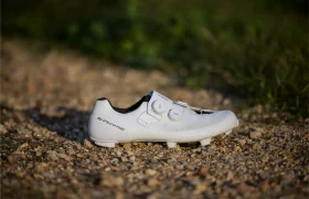 Una zapatilla de ciclismo RX910 S-Phyre blanca con dos esferas se encuentra en un camino de grava, rodeada de hierba y vegetación borrosa al fondo.