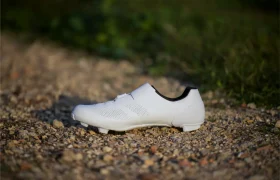 Unas Zapatillas Shimano RX910 S-Phyre blancas de ciclismo descansan sobre un camino de grava con vegetación en el fondo borroso.