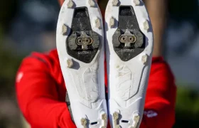 Una persona con chaqueta y casco rojos sostiene un par de zapatillas blancas de ciclismo Gravel con tacos SPD, con las suelas mirando a la cámara.