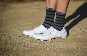 Persona de pie en un camino de grava con zapatillas de ciclismo Shimano RX910 S-Phyre blancas con dos cierres de esfera y calcetines a rayas negras y grises.
