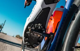 Primer plano de la zapatilla de un ciclista enganchada en LOOK pedales para Gravel, con un cuadro de bicicleta azul y un camino de tierra de fondo bajo un cielo despejado, captando la esencia de la unión de ciclista y bicicleta en el ecosistema de pedales.