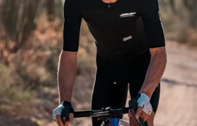 Ciclista con una equipación negra, casco blanco y gafas de sol naranjas monta en una bicicleta de grava con pedales LOOK para Gravel en un camino de tierra bordeado de cactus.