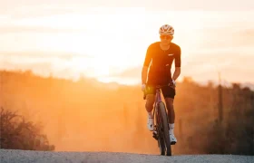 Ciclista con atuendo negro y casco monta una bicicleta con pedales LOOK para Gravel en un camino de grava al atardecer, la cálida luz del sol creando un fondo resplandeciente.