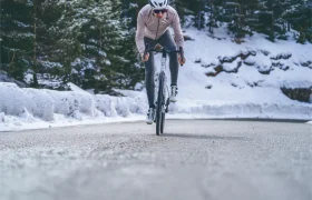 Un ciclista con casco y una Chaqueta Sportful Fiandre Shift monta en bicicleta por una carretera de montaña nevada y rodeada de árboles, disfrutando de protección y transpirabilidad.