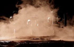 Tres objetos metálicos, como grava pedregosa, parcialmente oscurecidos por un espeso polvo o humo de color anaranjado, se yerguen sobre una superficie rocosa y desigual bajo una tenue iluminación.