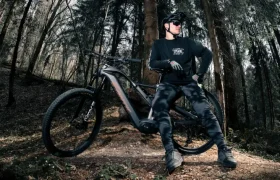 Persona con ropa de ciclismo y casco negros junto a una Fantic Sierra e-MTB, con motor Bosch CX Gen5, en una zona boscosa con árboles altos y el suelo cubierto de hojas.