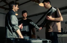 En el taller industrial, bajo la intensa luz de la iluminación cenital, tres personas examinan cuidadosamente un componente grande y oscuro. Una persona lo sostiene en alto —una pieza crucial para un proyecto UNNO MITH— mientras las demás observan con atención.