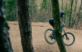 Ataviado con elegante equipo de ciclismo negro, un ciclista que luce la UNNO MITH se sube con destreza a una bicicleta de montaña por un sendero de tierra. El exuberante bosque de fondo, con sus imponentes árboles y su vibrante follaje, realza la emoción del paseo.