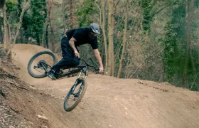 Una persona que lleva casco conduce una bicicleta de montaña UNNO MITH por una rampa de tierra en una zona boscosa.