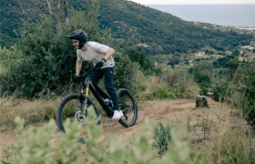 Un ciclista con casco maniobra con destreza una bicicleta de montaña UNNO MITH a lo largo de un sinuoso sendero de tierra, rodeado de exuberantes colinas verdes con el mar sereno extendiéndose más allá.
