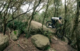 Una persona con casco conduce hábilmente una bicicleta de montaña UNNO MITH por una gran roca en una zona boscosa.