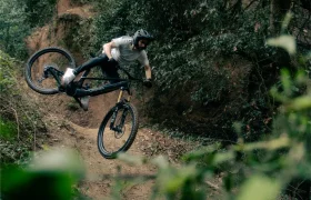 Un ciclista de montaña en un UNNO MITH se eleva por los aires, ejecutando un salto en un sendero de tierra, envuelto de exuberantes árboles y follaje.