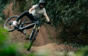 Un ciclista de montaña con una camisa blanca realiza hábilmente un truco en el sendero, levantando la rueda trasera mientras conduce por el área boscosa con su bicicleta UNNO MITH.