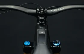 Primer plano de un manillar y una potencia de bicicleta UNNO MITH con empuñaduras negras y una pequeña pantalla en el centro, sobre un fondo oscuro.