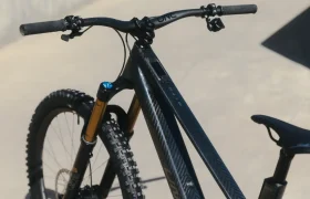 Primer plano de una elegante bicicleta de montaña UNNO MITH con cuadro texturizado, manillar ancho y suspensión delantera. La bicicleta negra brilla sobre una superficie de hormigón bajo la intensa luz del sol.